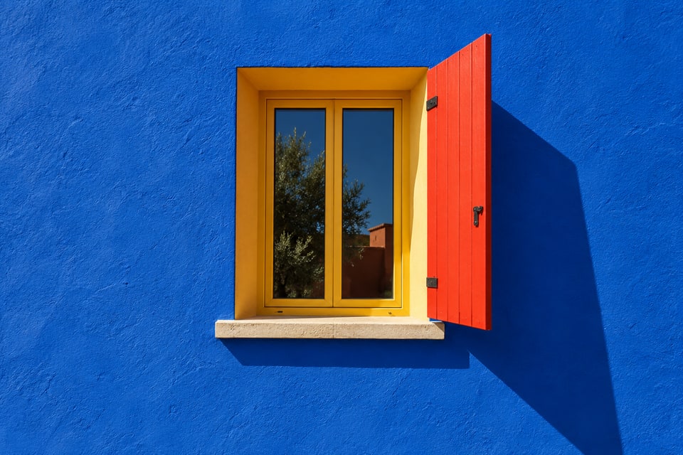 A marigold window with a red shutter in a cobalt wall