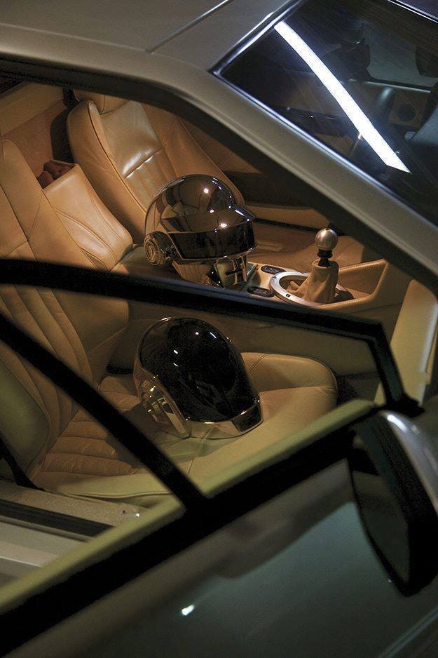 Two robot helmets, one chrome and one glossy black, resting on the tan leather seats of a car at night, seen through the window.