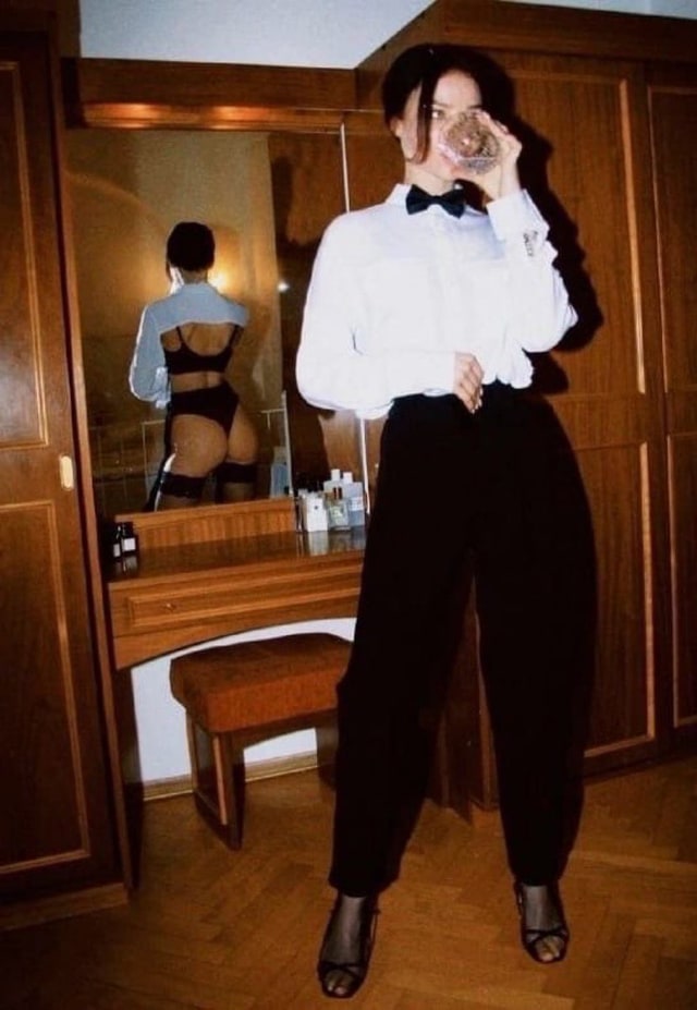 A woman in a tuxedo shirt before a vintage vanity mirror in warm tungsten light.