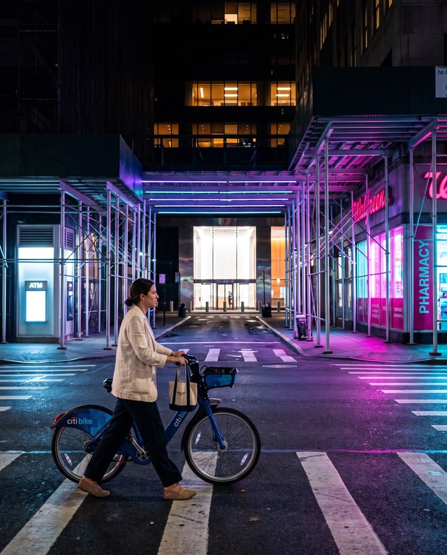 A woman walking a bike across a neon-lit crosswalk, made by Muse.